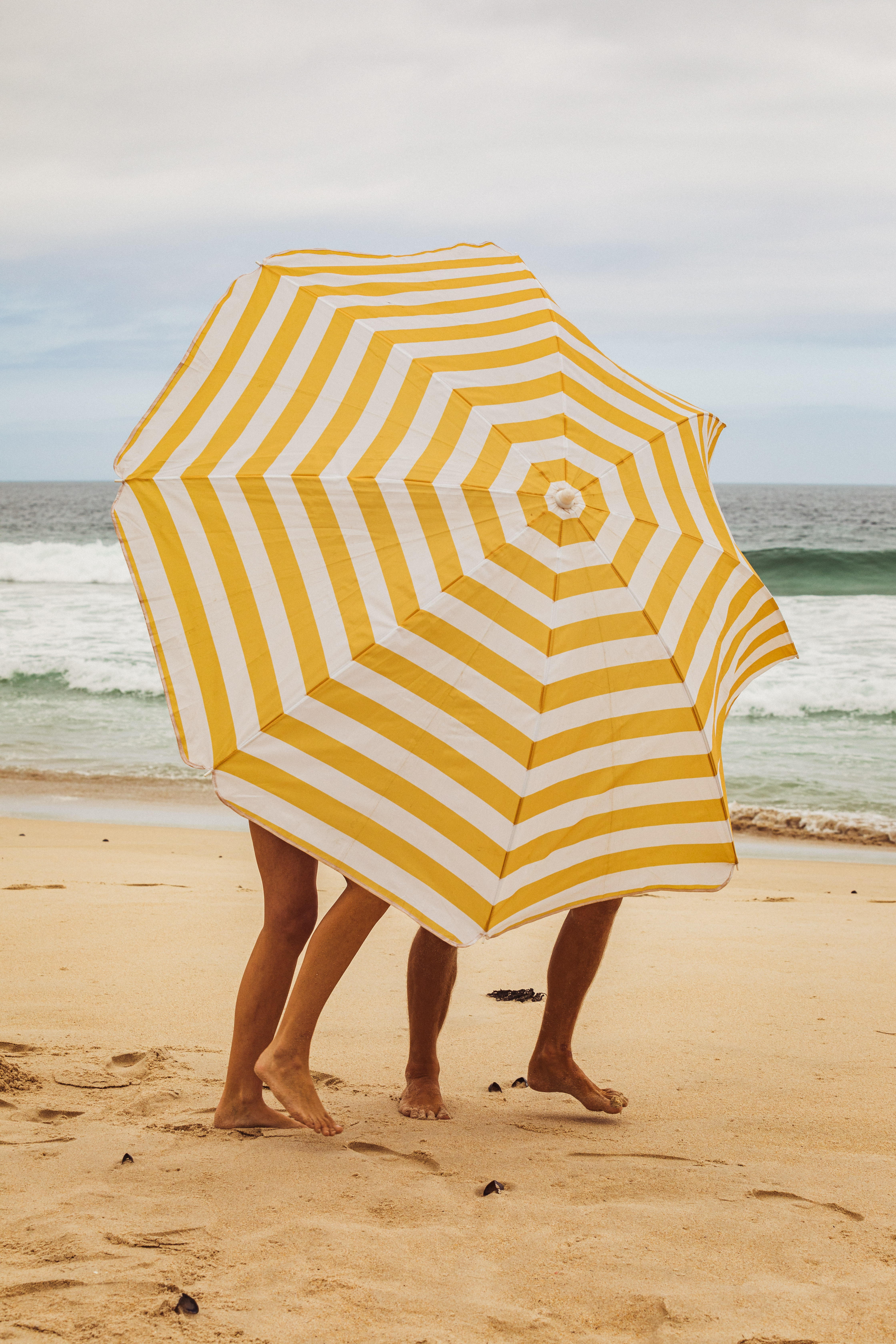 Yellow-and-white-striped parasol on the beach with the sea behind. In the background, waves roll onto the sandy beach, while the parasol fills most of the picture.