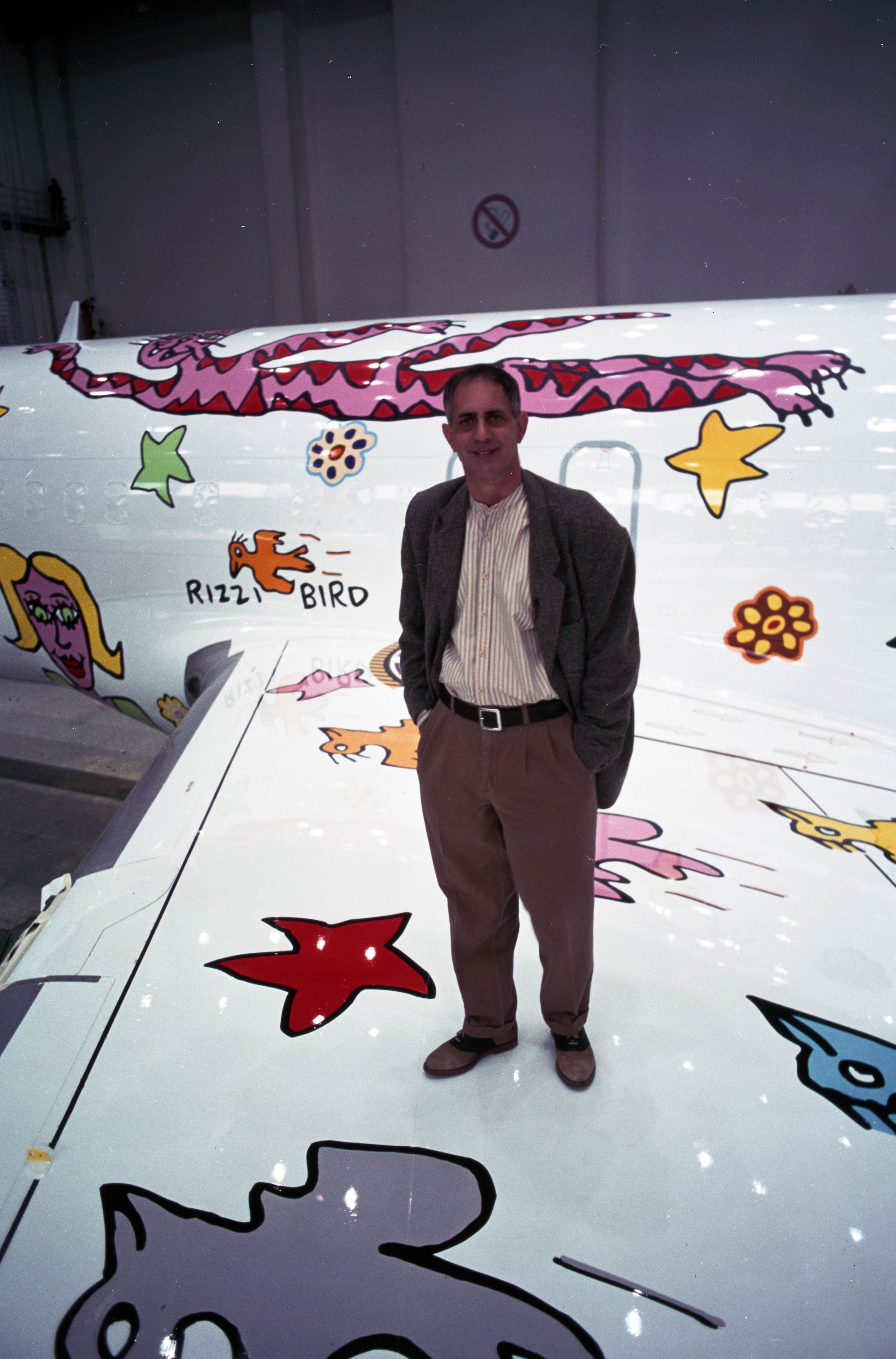 The picture shows a person in front of a passenger plane with special paintwork featuring colorful drawings, figures and stars. The person is standing in front of the aircraft fuselage and one wing. The photo was taken in an aircraft hangar.