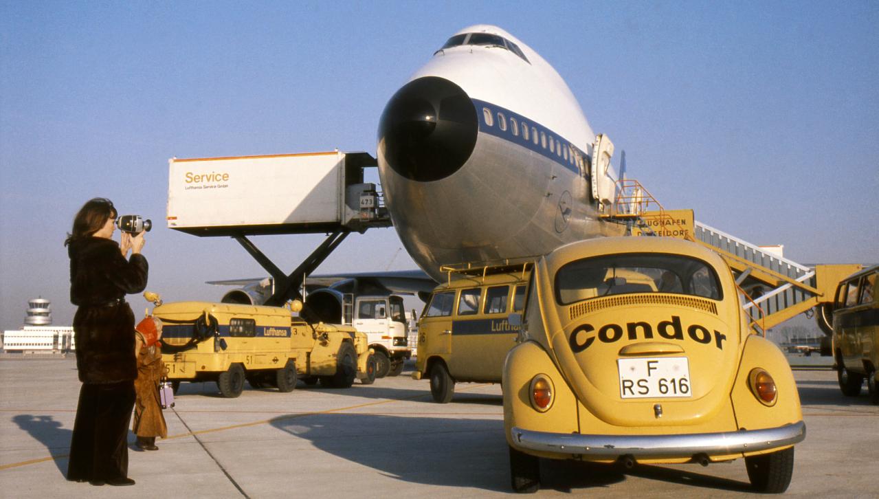 The picture shows the nose of a vintage passenger aircraft on an airport apron. In the foreground is a yellow VW Beetle with 'Condor' lettering. Next to the aircraft, other airport vehicles, a passenger staircase and two people can be seen, one of whom is a woman holding a camera. The aircraft is shown at an angle from the front.
