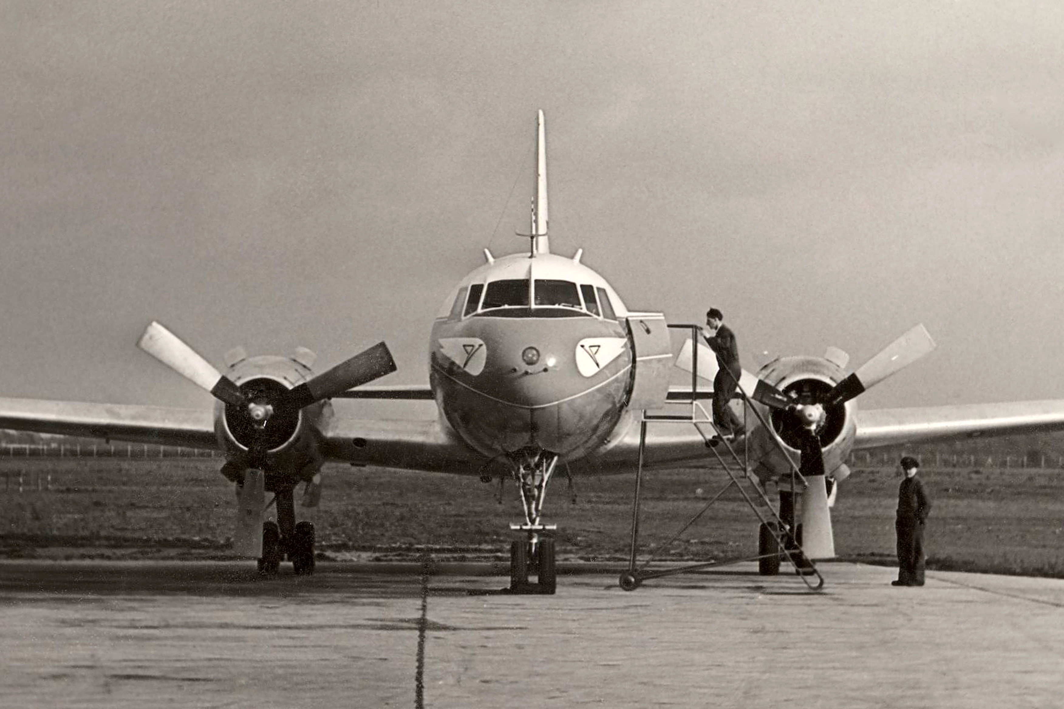 La photo montre un embarquement en 1958 au moyen d'un escalier mobile dans un Convair CV-240. Cet avion de ligne historique à hélices est vu de face sur le tarmac d'un aéroport. Une personne se tient sur l'escalier, au niveau de la porte ouverte de l'avion, tandis qu'une autre se tient à côté de l'appareil. La photo est en noir et blanc.