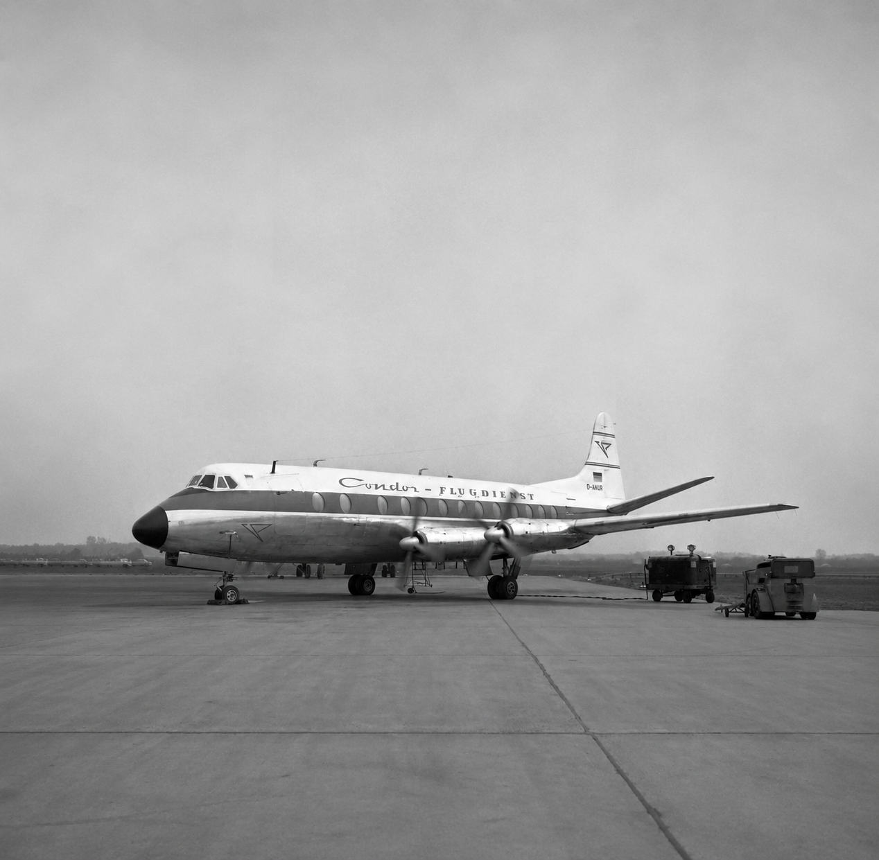 The picture shows a historic propeller-powered passenger plane on the apron of an airport. The aircraft is shown in profile. Several ground vehicles are next to the aircraft. The photograph is in black and white.