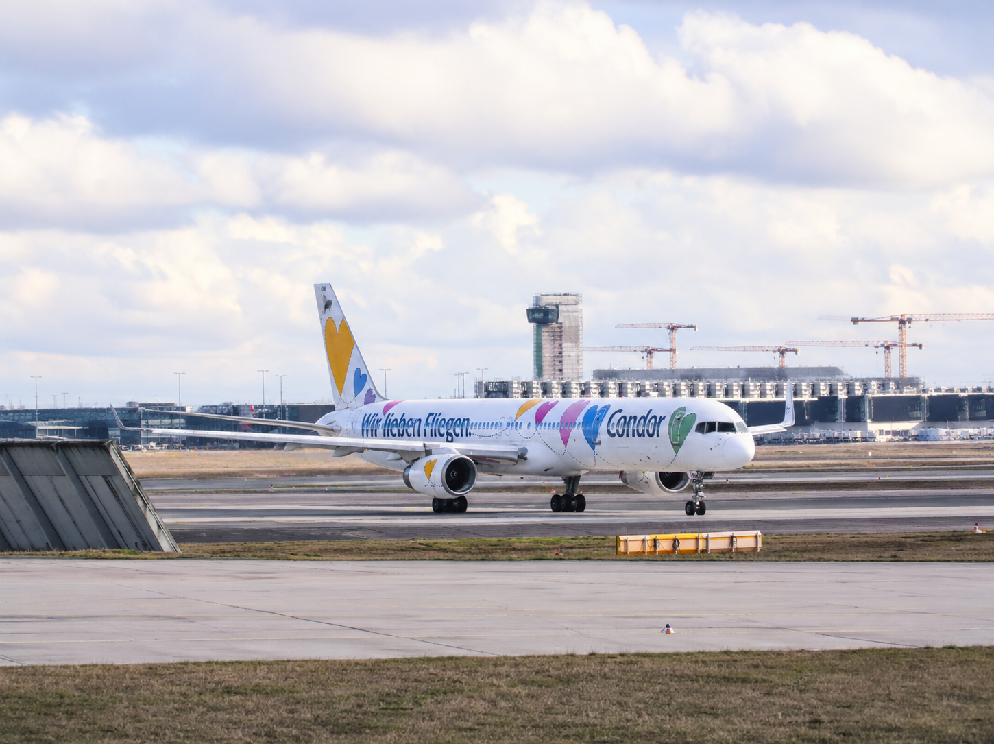 The picture shows a passenger plane with special paintwork on an airport runway. The aircraft fuselage features colorful heart motifs and the lettering “We love flying.” The aircraft is shown in profile. In the background, airport buildings, a control tower and construction cranes can be seen.
