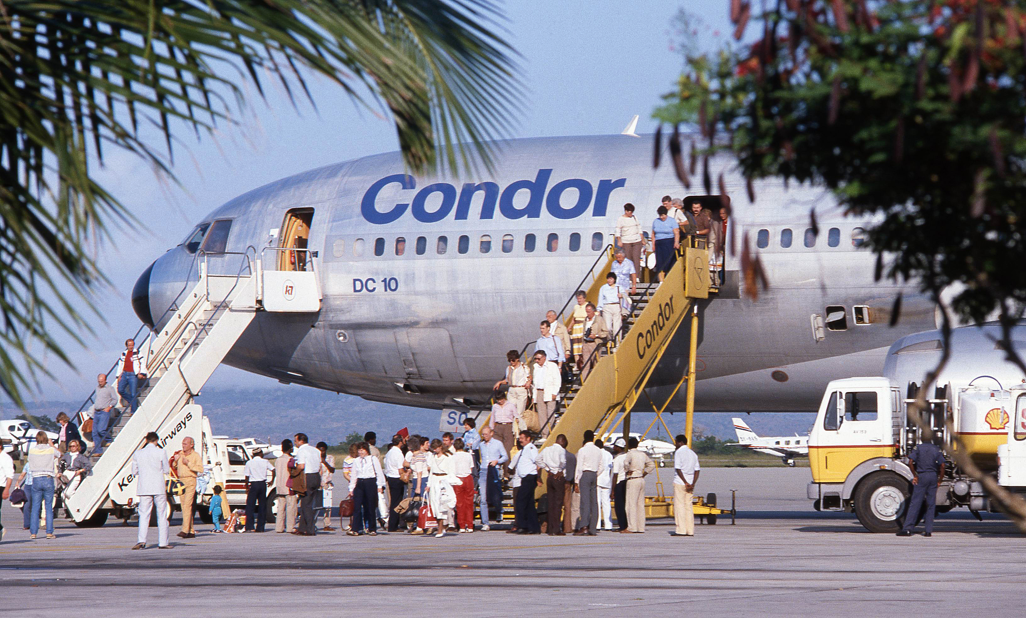 The picture shows passengers boarding and disembarking from a vintage passenger aircraft via two mobile staircases. There are several people on the steps and on the apron in front of the aeroplane. The aircraft is shown in profile. In the foreground, airport vehicles can be seen, while palm leaves frame the picture.