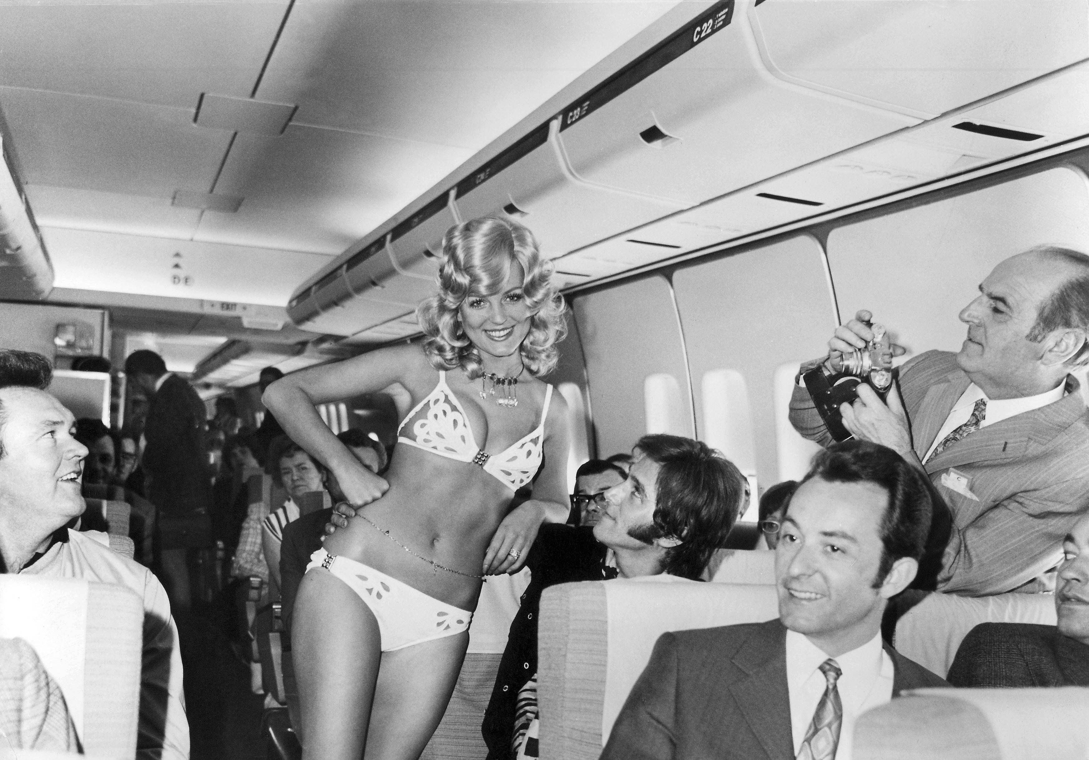The picture shows a woman in underwear standing in the cabin of a passenger plane between seated passengers. Several passengers look at the woman while another person takes pictures in the background. The photo is in black and white.