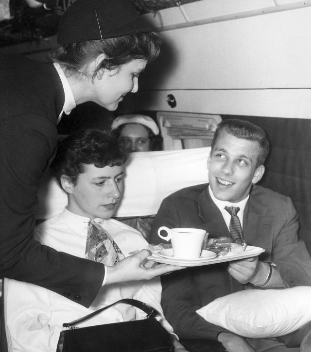 The picture shows a stewardess handing out a tray of drinks and food to two seated passengers in an aircraft cabin. The photo is in black and white. In the background, other passengers and the interior of the cabin can be seen.