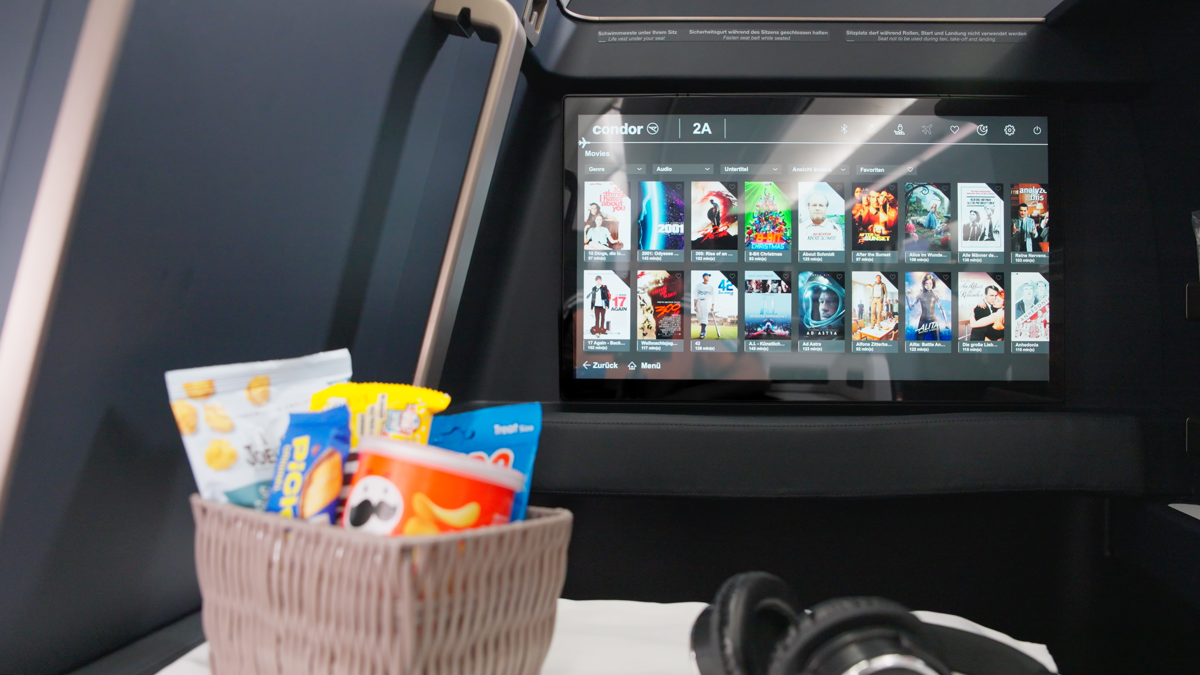 The picture shows a screen with the in-flight entertainment program in the backrest of an aircraft seat. Various movies are displayed on the screen. In the foreground are a basket of snacks and a pair of headphones. This was taken in the cabin of a passenger aircraft.