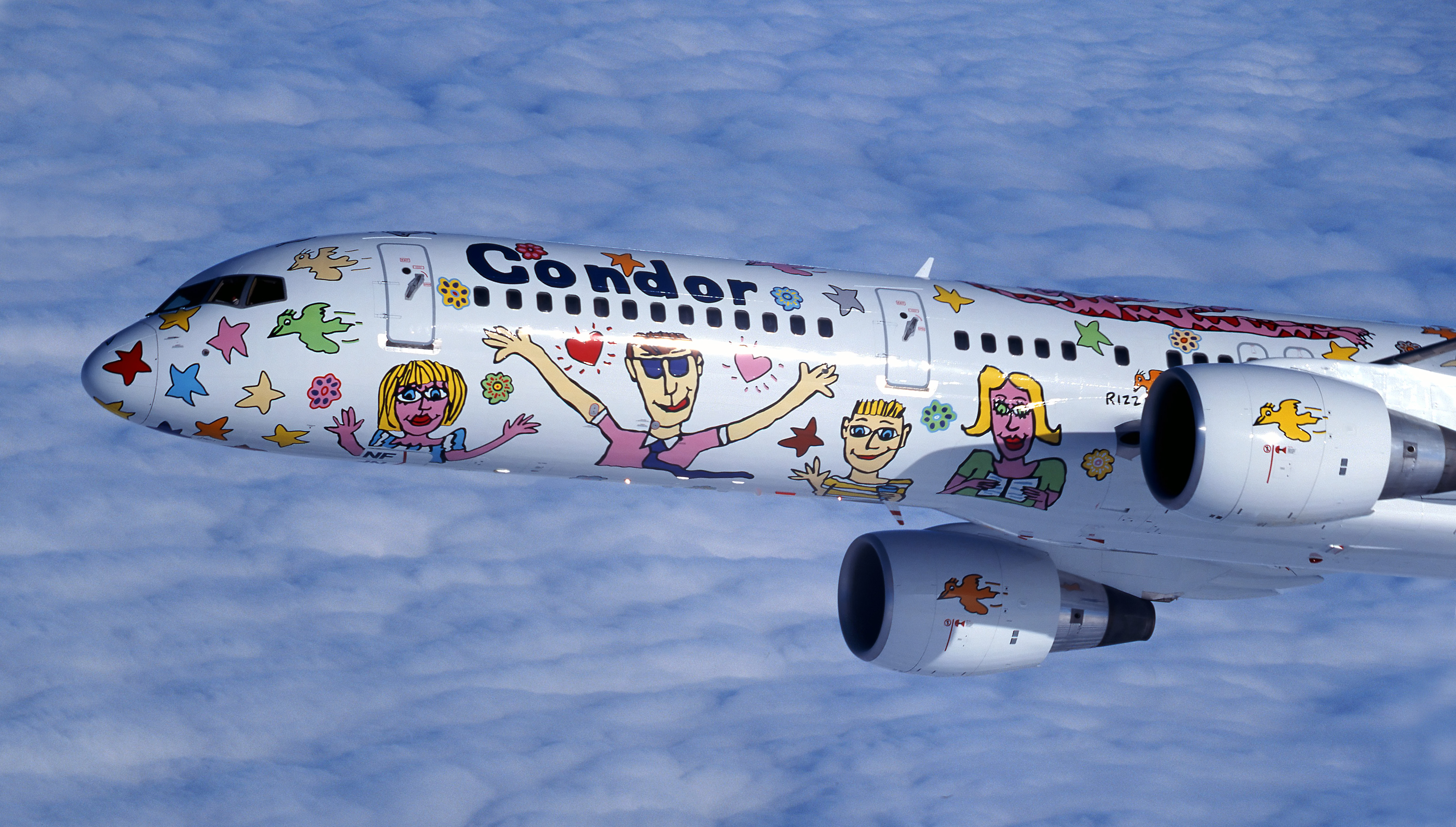 The picture shows a passenger plane with a special paint of colorful drawings, figures and stars during flight. The aircraft is shown at an angle from the side and flies over a solid cloud layer. In view are the front part of the fuselage and the engines.