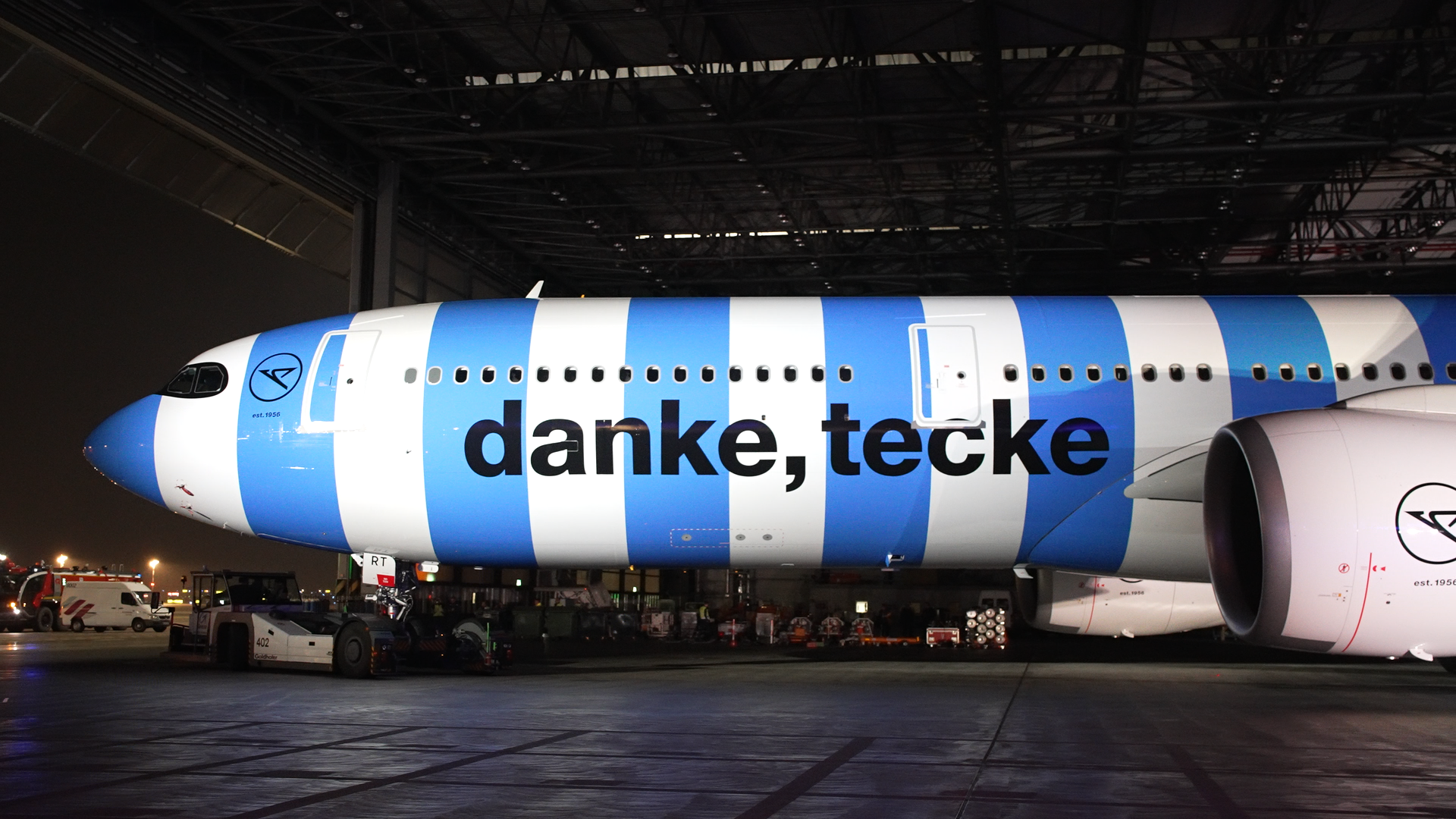 The picture shows the fuselage of a passenger aircraft in blue-white striped special livery in an aircraft hangar. The aircraft fuselage features the lettering “danke, tecke”. The aircraft is shown in profile.