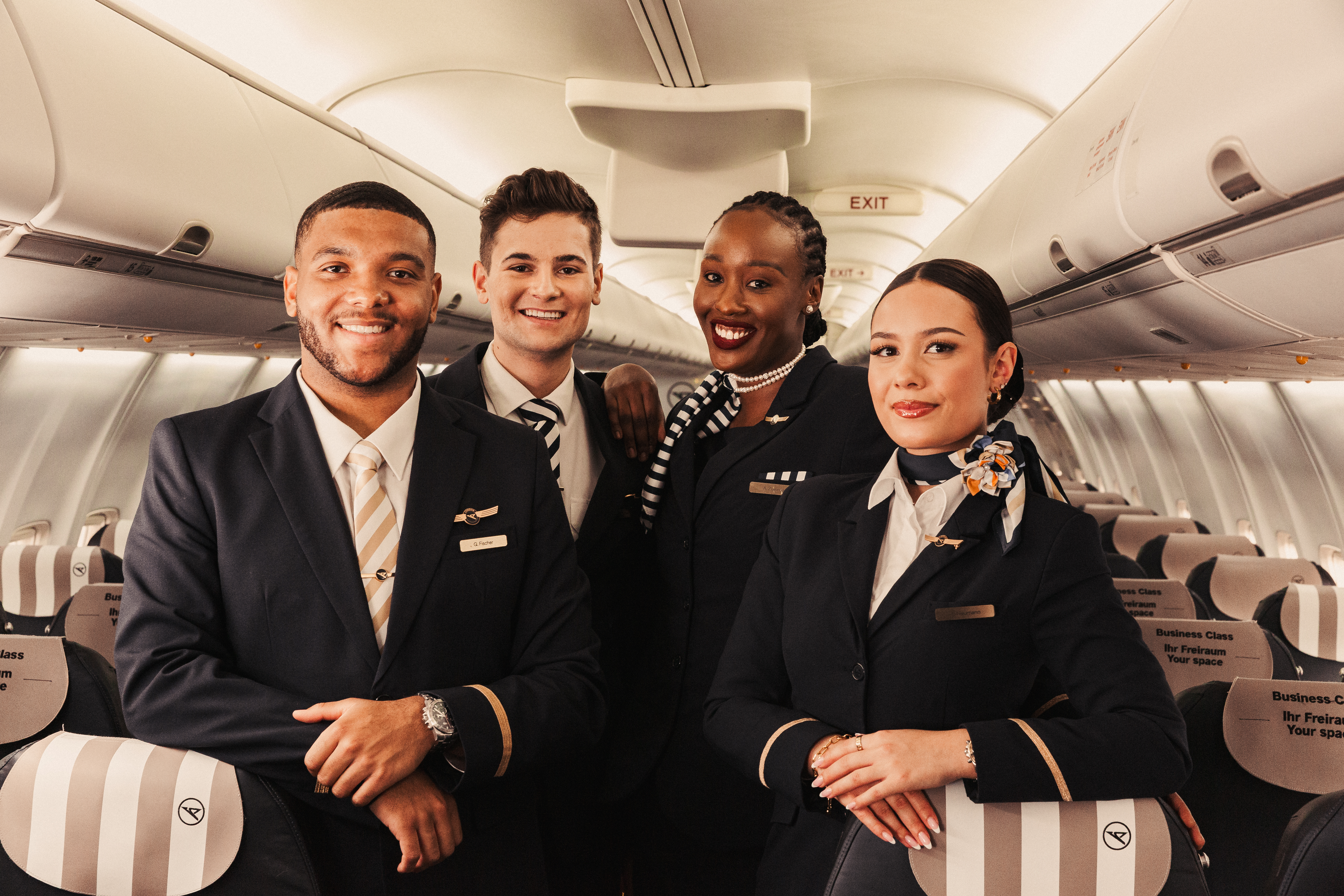 The image shows four crew members in dark uniforms in the cabin of a passenger plane. The people are standing next to each other in the aisle and looking into the camera. The cabin ceiling, overhead bins and rows of seats can be seen in the background.