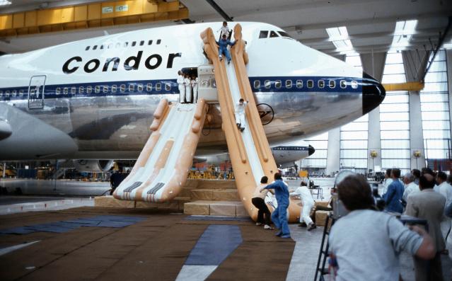 The picture shows the nose of a vintage passenger aircraft in a hangar. Inflated emergency slides are attached to the aircraft doors, on which several people are sliding down or standing. Other people are in front of the emergency slides. In the background, parts of other aircraft and the hangar facilities can be seen.